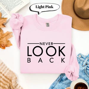 Pode incluir: Camisola rosa claro com a frase "NEVER LOOK BACK" em preto. A camisola está dobrada, com as mangas visíveis. A imagem também mostra um chapéu, calças de ganga e uma chávena de café.