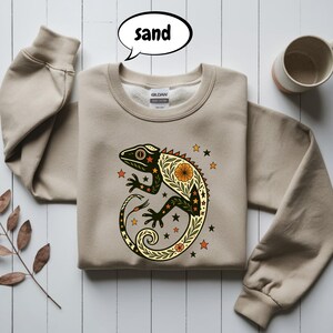 May include: A sand-coloured sweatshirt with a detailed lizard design. The lizard is black and green with orange and yellow floral patterns and star accents. The word "sand" is in a speech bubble.