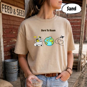 May include: Sand-colored t-shirt with the phrase "Born to Roam" and a graphic of a car, globe, airplane, and heart-shaped flight path. The shirt is a neutral color and has short sleeves.