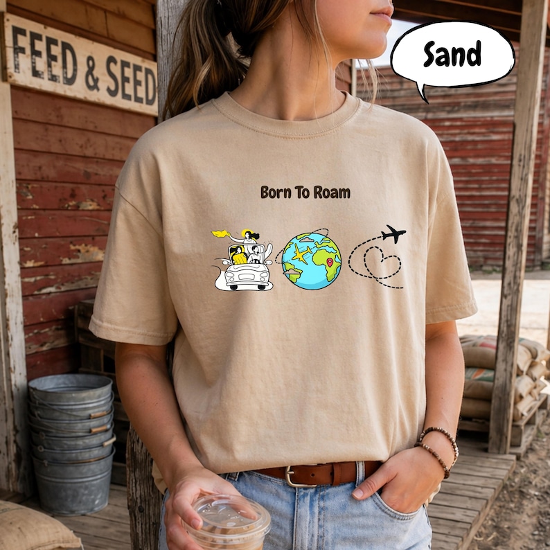 May include: Sand-colored t-shirt with the phrase "Born to Roam" and a graphic of a car, globe, airplane, and heart-shaped flight path. The shirt is a neutral color and has short sleeves.