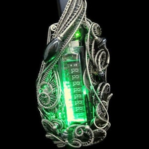 Puede incluir: Un colgante de alambre plateado con una pantalla digital verde brillante en el interior. El colgante está en un cordón negro.