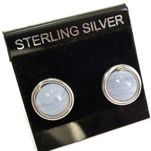 Pode incluir: Um par de brincos de pressão com pedras redondas azuis claras emolduradas em metal prateado. Os brincos são exibidos em um cartão preto com o texto "STERLING SILVER".