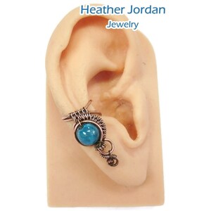 May include: A copper wire ear cuff with a turquoise gemstone. The cuff is designed to wrap around the upper part of the ear.