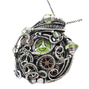 May include: Steampunk-style pendant necklace with intricate wire-wrapped design. The silver-toned pendant features gears, green gemstones, and a central green-tinted glass element. The necklace hangs from a delicate silver chain.