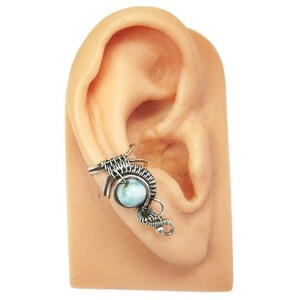 May include: A silver ear cuff with a blue gemstone. The cuff is made of wire and has a delicate design. It is worn on a model's ear.