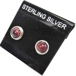 Pode incluir: Um par de brincos de prata esterlina com pedras de olho de tigre vermelho. Os brincos estão em um cartão preto com as palavras "Sterling Silver" impressas nele.