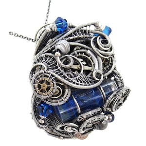 May include: A steampunk-style pendant necklace with a silver wire-wrapped design. The pendant features a rectangular blue crystal, small gears, and blue crystal accents. The necklace has a silver chain.