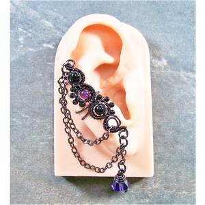 May include: A dark metal ear cuff featuring a decorative design with black and purple gemstones. The cuff has a chain that dangles down with a purple teardrop-shaped crystal at the end. The ear cuff is displayed on a mannequin ear.