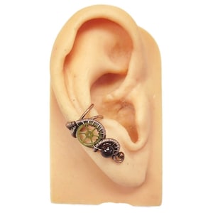May include: A copper and green ear cuff with a gear design and a black bead. The cuff is worn on a model ear.
