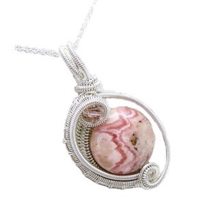Pode incluir: Pingente artesanal com uma pedra preciosa redonda rosa e branca, envolta em fio de prata. O pingente pende de uma delicada corrente de prata, mostrando o intrincado trabalho de arame e os padrões únicos da pedra natural.