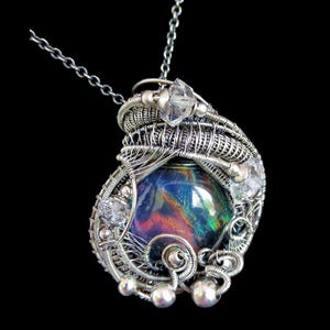 Puede incluir: Un collar con colgante de plata hecho a mano con una gran piedra preciosa iridiscente en el centro. La piedra preciosa muestra un espectro de colores. El colgante está intrincadamente envuelto en alambre con una delicada cadena.