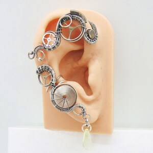 May include: Silver ear cuff with a steampunk design featuring gears and a dangling green stone.