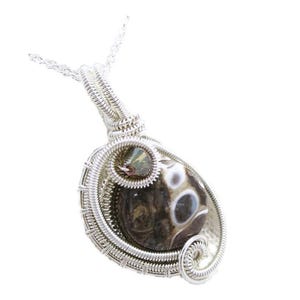 Pode incluir: Pingente artesanal com uma pedra polida em forma oval, com um padrão único em marrom, branco e cinza. A pedra é envolvida em um fio de prata detalhado, suspensa por uma delicada corrente de prata. O pingente tem um design em espiral na parte inferior.