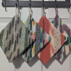 Puede incluir: Cuatro agarraderas cuadradas hechas a mano de varios colores cuelgan de un estante de metal negro. Las agarraderas están hechas de hilo en tonos verdes, grises, rosas y azules. Cada agarradera tiene un lazo para colgar.