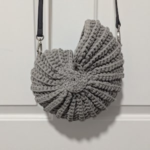 May include: A gray, handmade crochet bag with a unique, seashell-like design. The bag features a long, black strap and silver-toned hardware. The bag's texture is visible, showcasing the intricate crochet work.