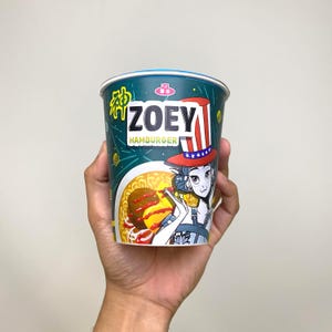 May include: A cup of Zoey Hamburger instant noodles held in a hand. The cup features a cartoon illustration of a woman wearing a patriotic hat. The cup has the words "Zoey Hamburger" printed on it, with a green and blue color scheme.