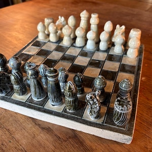 Puede incluir: Un juego de ajedrez de cerámica con un tablero a cuadros blanco y negro. Las piezas del ajedrez son de dos colores: blanco roto y verde oscuro. El juego de ajedrez está sobre una superficie de madera.