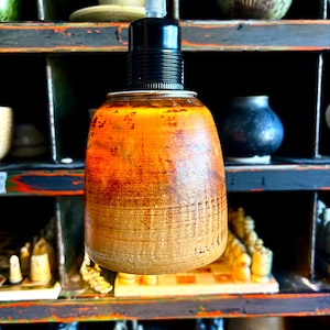 May include: A handcrafted, ceramic pendant light fixture with a warm, earthy color gradient. The fixture has a black socket and cord, and is suspended against a backdrop of shelves with various decorative items.