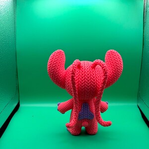 Peut inclure: Une poup&eacute;e Stitch rose tricot&eacute;e &agrave; la main, avec de grandes oreilles, des accents violets et un triangle bleu sur le dos. La poup&eacute;e est devant un fond vert.