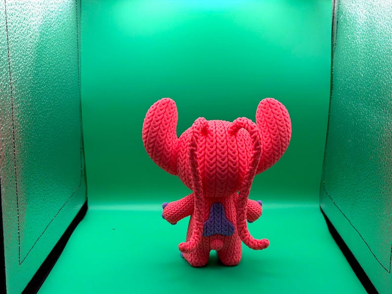 Peut inclure: Une poup&eacute;e Stitch rose tricot&eacute;e &agrave; la main, avec de grandes oreilles, des accents violets et un triangle bleu sur le dos. La poup&eacute;e est devant un fond vert.