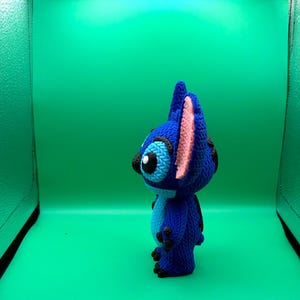 May include: A blue crocheted Stitch figure with black eyes, ears, and feet, and pink inner ears. The figure is standing against a green background.