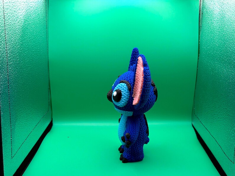 May include: A blue crocheted Stitch figure with black eyes, ears, and feet, and pink inner ears. The figure is standing against a green background.
