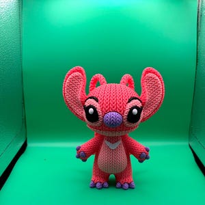 Peut inclure: Un personnage crochet&eacute; rose et violet fait &agrave; la main, avec de grandes oreilles, des yeux noirs et un nez violet. Le personnage a un chevron blanc sur sa poitrine et des pieds et des mains violets. Le fond est vert uni.