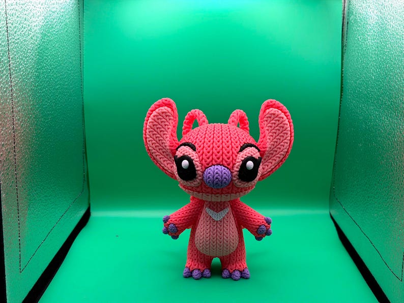 Peut inclure: Un personnage crochet&eacute; rose et violet fait &agrave; la main, avec de grandes oreilles, des yeux noirs et un nez violet. Le personnage a un chevron blanc sur sa poitrine et des pieds et des mains violets. Le fond est vert uni.