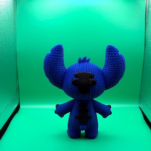 May include: A blue knitted Stitch doll with large ears, black eyes, and a black patch on its chest. The doll is standing against a green background.