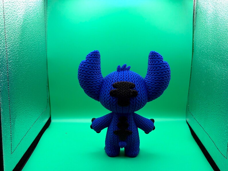 May include: A blue knitted Stitch doll with large ears, black eyes, and a black patch on its chest. The doll is standing against a green background.