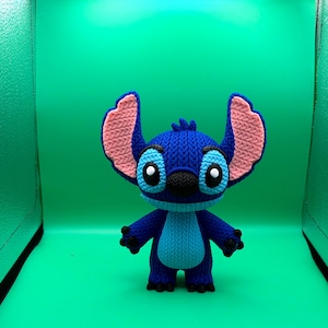 May include: A blue and pink crocheted Stitch figure, featuring large ears, black eyes, and a black nose. The figure is standing against a bright green background.