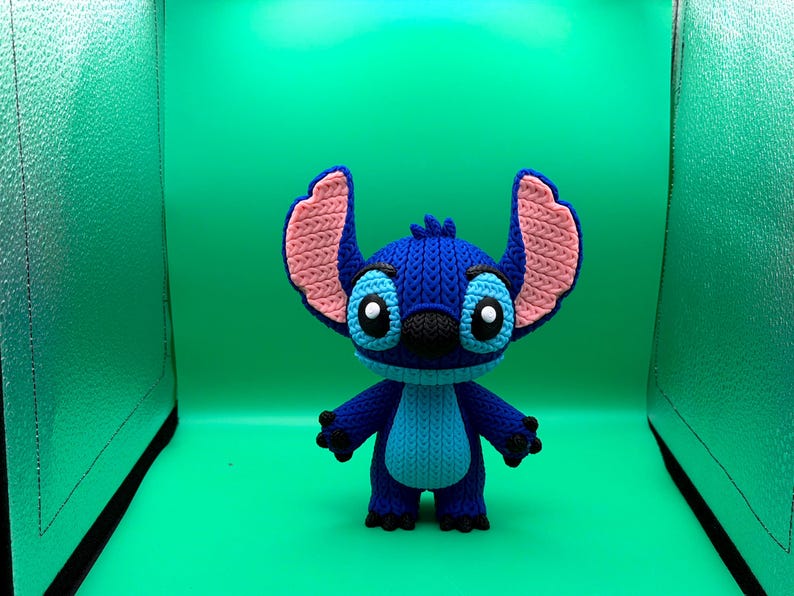 May include: A blue and pink crocheted Stitch figure, featuring large ears, black eyes, and a black nose. The figure is standing against a bright green background.