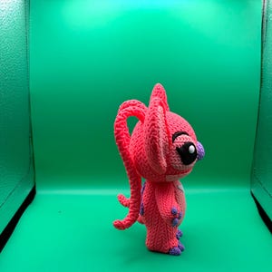 Peut inclure: Une figurine rose au crochet faite &agrave; la main, avec de grandes oreilles et une longue queue boucl&eacute;e. La figurine a des yeux noirs, un nez violet et des accents violets sur les mains et les pieds. Le fond est vert uni.