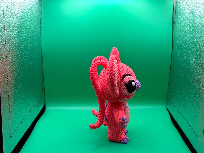 Peut inclure: Une figurine rose au crochet faite &agrave; la main, avec de grandes oreilles et une longue queue boucl&eacute;e. La figurine a des yeux noirs, un nez violet et des accents violets sur les mains et les pieds. Le fond est vert uni.