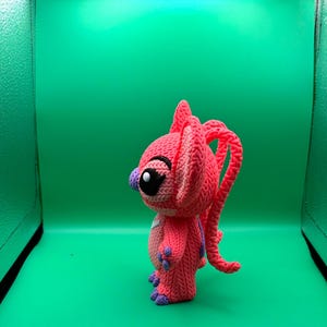 Peut inclure: Poup&eacute;e rose au crochet faite &agrave; la main, avec de grands yeux noirs, des accents violets et de longs cheveux boucl&eacute;s. La poup&eacute;e a une apparence textur&eacute;e et d&eacute;taill&eacute;e et se tient sur un fond vert. Le design de la poup&eacute;e rappelle un personnage de dessin anim&eacute;.