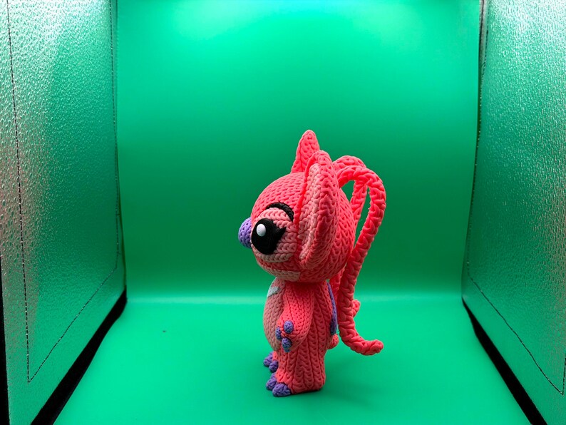 Peut inclure: Poup&eacute;e rose au crochet faite &agrave; la main, avec de grands yeux noirs, des accents violets et de longs cheveux boucl&eacute;s. La poup&eacute;e a une apparence textur&eacute;e et d&eacute;taill&eacute;e et se tient sur un fond vert. Le design de la poup&eacute;e rappelle un personnage de dessin anim&eacute;.