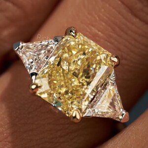 May include: A three-stone ring featuring a large, rectangular radiant-cut yellow center stone held by four prongs, flanked by two clear, triangular-cut side stones set in a metallic band. The design showcases a prominent yellow gemstone.