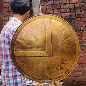 Könnte beinhalten: Ein runder Schild im antiken Stil mit Bronze-Finish. Der Schild hat ein markantes L-förmiges Design in der Mitte und eine strukturierte Oberfläche. Der Schild wird vor einer Person mit einem karierten Hemd gehalten.