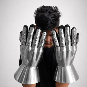 May include: A pair of articulated metal gauntlets featuring polished silver-colored steel plates covering the fingers and back of the hand, attached to a black fabric glove base with a flared cuff design at the wrist for protective coverage.