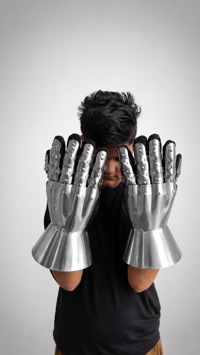 May include: A pair of articulated metal gauntlets featuring polished silver-colored steel plates covering the fingers and back of the hand, attached to a black fabric glove base with a flared cuff design at the wrist for protective coverage.