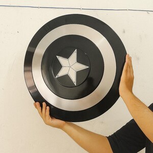 Op de afbeelding: Een zwart en zilver cirkelvormig schild met een witte ster in het midden. Het schild heeft een glanzende afwerking en wordt tegen een witte muur gehouden. Een rekwisiet of verzamelobject.