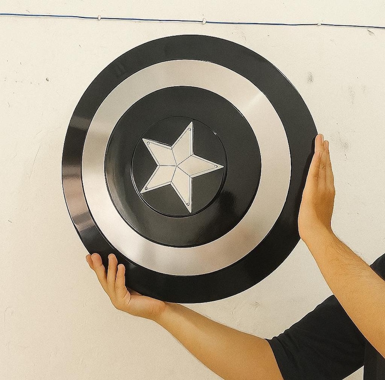 Op de afbeelding: Een zwart en zilver cirkelvormig schild met een witte ster in het midden. Het schild heeft een glanzende afwerking en wordt tegen een witte muur gehouden. Een rekwisiet of verzamelobject.