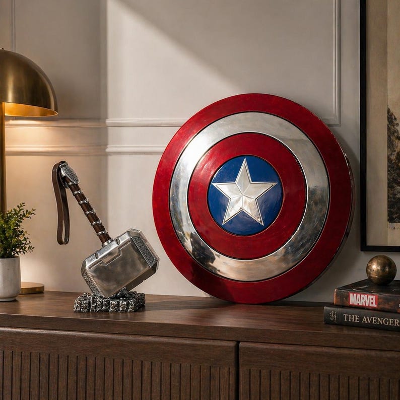Puede incluir: Un escudo del Capit&aacute;n Am&eacute;rica rojo, blanco y azul, y un martillo de Thor plateado con un mango marr&oacute;n. El escudo presenta una estrella blanca sobre un c&iacute;rculo azul. El martillo descansa sobre una base decorativa. Decoraci&oacute;n tem&aacute;tica de Marvel.