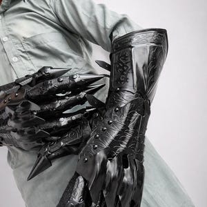 May include: A pair of black articulated gauntlets featuring long, pointed claw-like fingertips and intricate embossed scrollwork patterns across the hand and forearm plates. The armor pieces are secured with rivets and adjustable straps along the wrist.