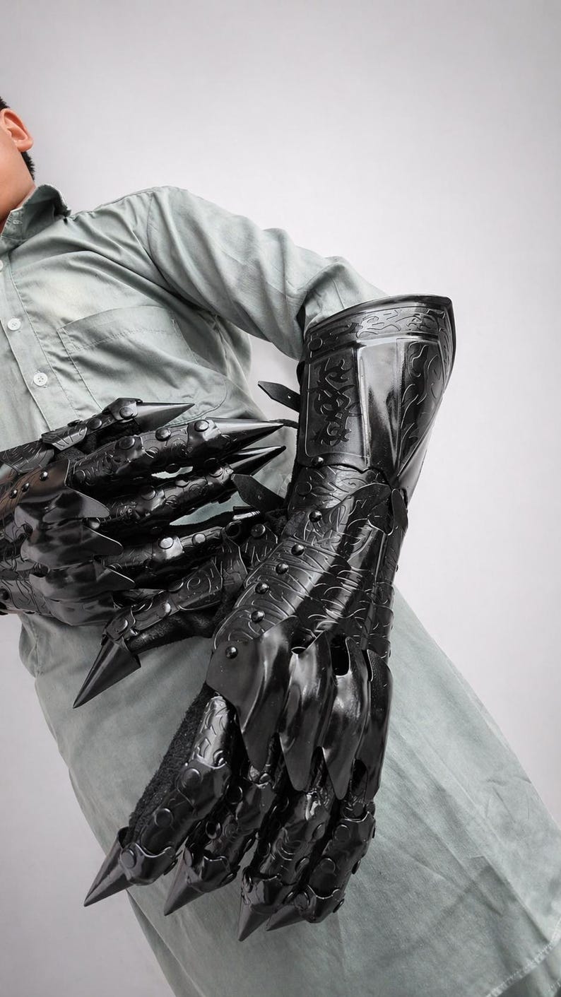 May include: A pair of black articulated gauntlets featuring long, pointed claw-like fingertips and intricate embossed scrollwork patterns across the hand and forearm plates. The armor pieces are secured with rivets and adjustable straps along the wrist.
