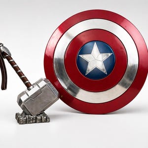 Puede incluir: Un escudo rojo, blanco y azul del Capit&aacute;n Am&eacute;rica con una estrella plateada en el centro, junto a un martillo plateado de Thor con un mango marr&oacute;n. El martillo descansa sobre una base texturizada y el escudo tiene un borde plateado.