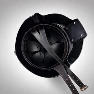 May include: A black metal helmet viewed from above, featuring a circular interior with a black leather suspension liner and a multi-pointed star-shaped crown pad. A long black leather chin strap with metal buckles extends from the helmet shell.