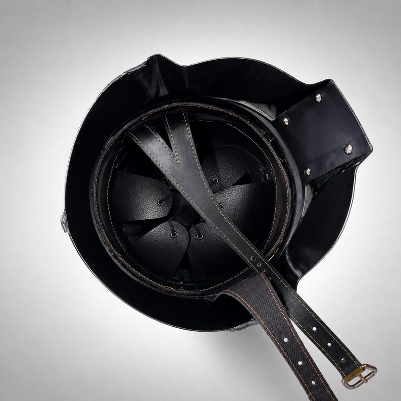 May include: A black metal helmet viewed from above, featuring a circular interior with a black leather suspension liner and a multi-pointed star-shaped crown pad. A long black leather chin strap with metal buckles extends from the helmet shell.