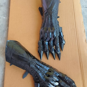 May include: A pair of black articulated gauntlets featuring a glossy finish, intricate embossed detailing on the forearm plates, and sharp, pointed finger extensions that mimic claw-like structures, designed to cover the hand and wrist area.