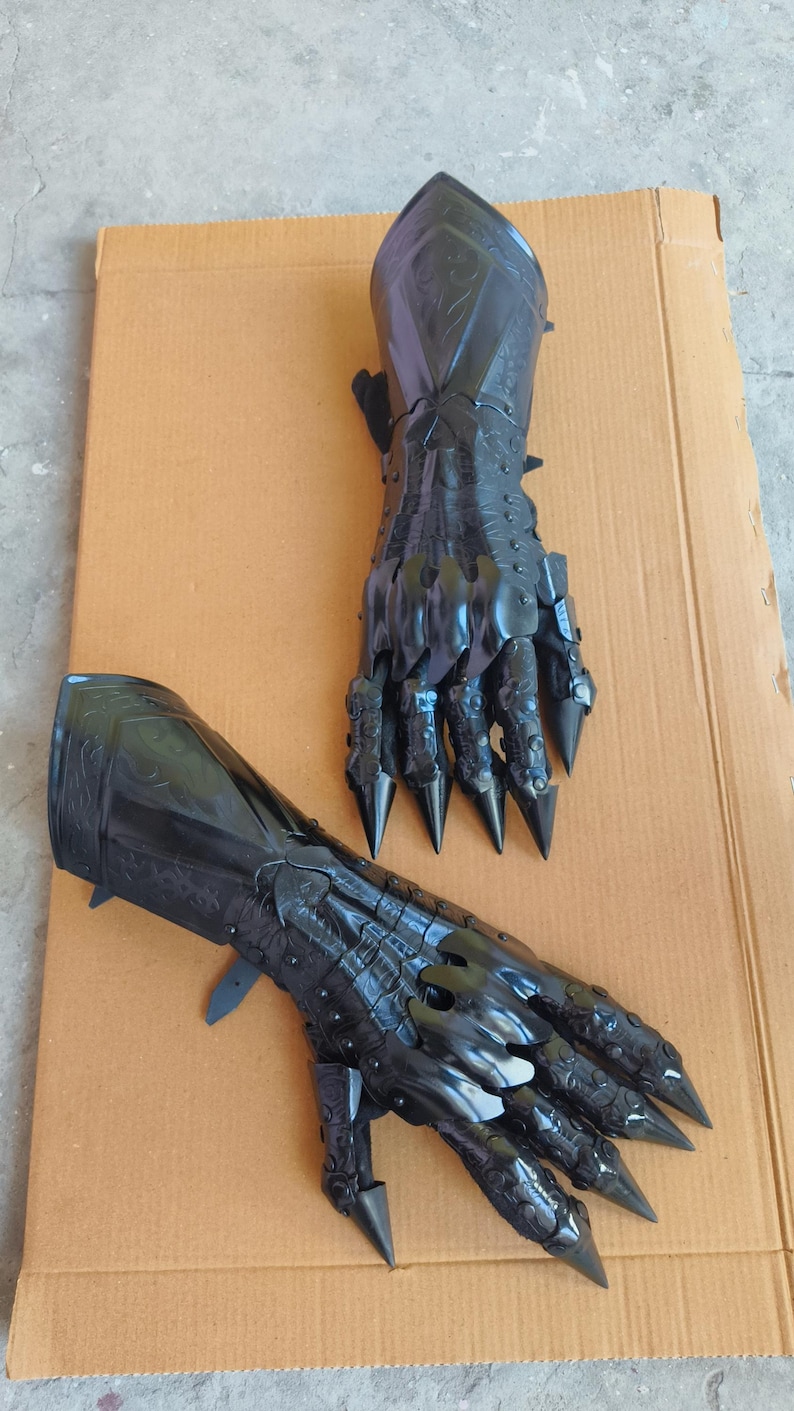 May include: A pair of black articulated gauntlets featuring a glossy finish, intricate embossed detailing on the forearm plates, and sharp, pointed finger extensions that mimic claw-like structures, designed to cover the hand and wrist area.